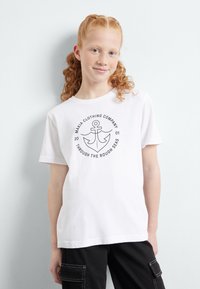 Camiseta de algodón blanca de manga corta, con un gráfico de ancla negra y texto en un diseño circular. El diseño incluye un tema náutico.