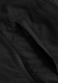 Svart tyg med en slät textur, som har en ficka med dragkedja och kontrasterande sömmar längs kanterna. Dragkedjan är i svart plast.