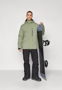 Roheline veekindel jakk eesmise lukuga, cargo taskute ja kapuutsiga. Mustad lumepüksid ja saapad. Inimene hoiab lumelauda.