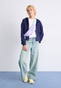 El modelo lleva una sudadera azul marino, una camiseta blanca, unos vaqueros de pierna ancha en azul claro y unas zapatillas amarillas, complementando el look con un bolso cruzado de un ligero color púrpura.