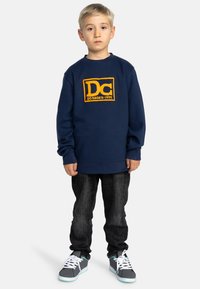 Navyblauwe sweatshirt met een oranje "DC" logo, gecombineerd met donkergrijze jeans en grijze sneakers met witte veters en turquoise accenten.