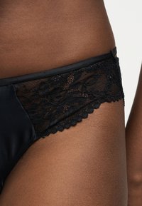 Μαύρα εσώρουχα δαντέλας με λουλουδένια σχέδια, φορεμένα σε σκούρο δέρμα, που δείχνουν την περιοχή των γοφών και του άνω μηρού σε απλό φόντο.