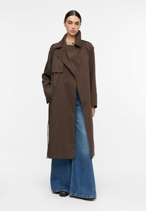 Frau steht und trägt einen langen dunkelbraunen Trenchcoat über weiten blauen Jeans und dunklen Schuhen vor einem schlichten weißen Hintergrund.
