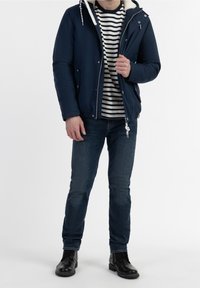 Homme portant une veste marine avec doublure sherpa blanche, chemise rayée noir et blanc, jean bleu et bottes en cuir noir, debout devant un fond blanc.