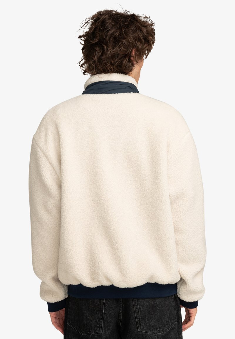 Chaqueta de forro polar color crema con una superficie suave y texturizada, que presenta un cuello y puños azul marino, dobladillo elástico y un corte holgado.