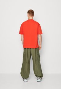 T-shirt en coton rouge à manches courtes, associé à un pantalon cargo ample vert olive avec des poches latérales, vu de l'arrière.