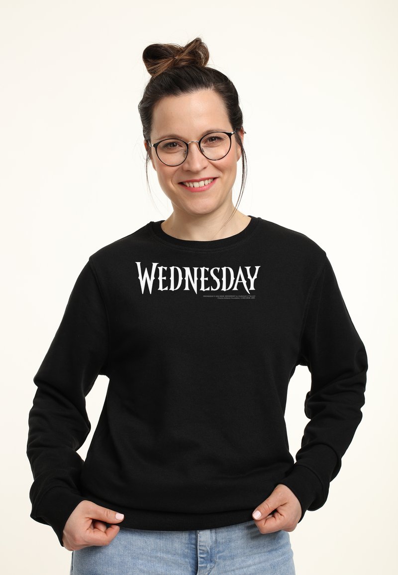 Donna sorridente con occhiali e capelli raccolti in un bun, indossa una felpa nera con la scritta "WEDNESDAY", mentre tiene l'orlo e si trova davanti a uno sfondo bianco.