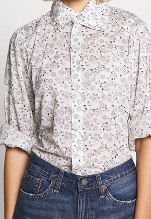 Personne portant une chemise blanche à boutons avec imprimé floral noir et manches retroussées, rentrée dans un jean en denim bleu.