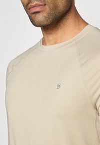 Beige színű, hosszú ujjú póló, kerek nyakkivágással, sima textúrával és egy kis logóval a bal oldalon. A varrási részletek láthatóak.