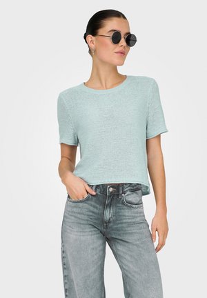 Ljusblått stickat kortärmat tröja med rund halsringning, ihop med bleknade gråa vida jeans. Svarta cirkulära solglasögon bärs.