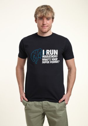 Sort t-shirt med hvid og blå tekst: "JEG LØBER MARATONER, HVAD ER DIN SUPERKRAFT?" med et grafisk motiv af en løber. Materiale: bomuld, rund halsudskæring.
