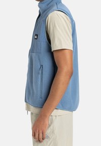 Modrá fleecová vesta s vysokým límcem, kapsou na hrudi a bočním zipem. Má texturovaný vnější povrch a hladkou vnitřní podšívku.