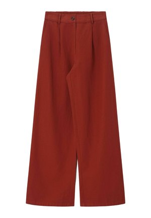 Femme aux longs cheveux ondulés porte des lunettes de soleil foncées, un haut à manches courtes et col en V de couleur rouge rouille, un pantalon large assorti et des chaussures beiges, mains dans les poches.