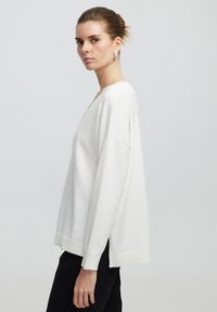 Bílý oversize svetr s V výstřihem a dlouhými rukávy, s žebrovanými manžetami a lemem, vyrobený z měkké, strukturované tkaniny. Viditelné jsou černé kalhoty.