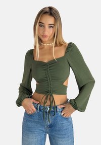Top verde oliva con maniche lunghe a sbuffo, parte anteriore arricciata, lati con tagli e dettagli con cordoncino. Abbinato a jeans blu a vita alta.