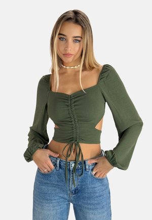 Olive green top with long puff sleeves, ruched front, cut-out sides, and drawstring detail. Paired with high-waisted blue jeans.