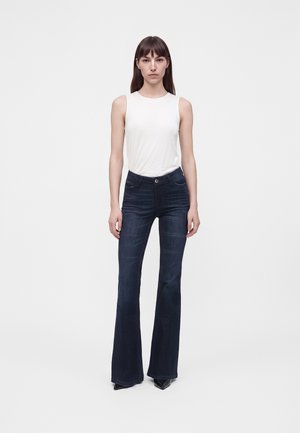 Woman standing facing forward wearing a white sleeveless top and dark blue flared jeans against a plain light background.