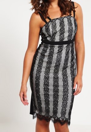 Frau trägt ein ärmelloses schwarz-weißes Kleid mit vertikalen schwarzen Spitzestreifen und einem Spitzenabschluss mit gewelltem Rand.