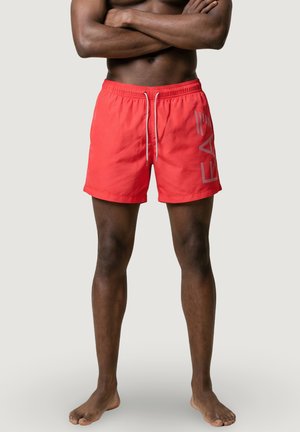 Uomo in piedi a piedi nudi con le braccia incrociate che indossa pantaloncini da bagno rossi vivaci con la scritta "LEA" sul lato, su uno sfondo chiaro e uniforme.