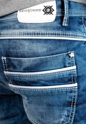 Blauwe denim jeans met meerdere zakken, contrasterende witte stiksels en een textuurlabel met zwarte branding zichtbaar op de tailleband.