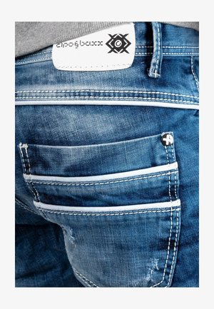 Jeans in denim blu con numerose tasche, cuciture bianche a contrasto e un'etichetta testurizzata con marchio nero visibile sulla vita.