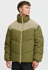 Chaqueta acolchada verde y beige con un alto cuello, cierre de cremallera frontal y un diseño de panel geométrico en contraste. Tejido suave con textura acolchada.