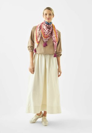 Frau stehend, trägt eine beige Bluse, einen langen weißen Rock, weiße Sneakers und einen bunten, gemusterten Schal mit Quasten um den Hals.