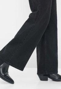 Jeans neri a gamba larga con una texture liscia, abbinati a stivali neri con punta e tacco basso a forma di blocco.