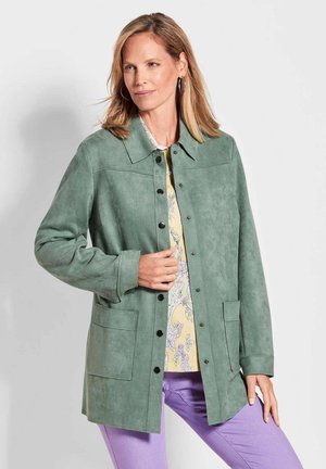 Groene suède jas met een kraag, knopen aan de voorkant en twee zakken. Draagt over een gemusterde gele top en gecombineerd met paarse broek.