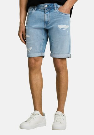 Mann mit hellblauen, hochgekrempelten, abgenutzten Jeansshorts und weißen Sneakers, stehend mit einer Hand in der Tasche.