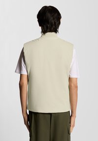 Ljusbeige ärmlös väst med hög krage och slät yta, bärs över en vit t-shirt. Enkelt design, inga synliga stängningar.