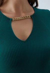 Donna che indossa un top teal a coste con un collo a chiave, collegato da una breve catena d'oro attraverso la parte superiore del petto.