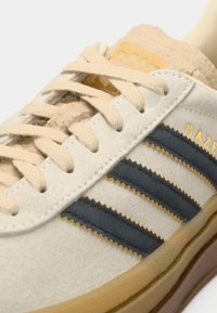 White suede sneaker with navy stripes, beige laces, and a textured tan sole. "GAZELLE" and "adidas" logos in gold on the side.