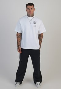 Balts īsām piedurknēm t-krekls ar logo, apvienots ar melniem platākām biksēm. Abi ir izgatavoti no kokvilnas. Pelēki ikdienas sporta apavi noslēdz apģērbu.