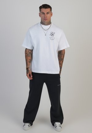 Fehér, rövid ujjú póló logóval, amelyet fekete, bő szárú nadrággal párosítottak. Mindkettő pamutból készült. A szettet szürke, sportos sneakers kiegészítik.