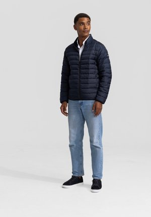 Marine blauwe pufferjack met een ritssluiting aan de voorkant, staande kraag en horizontaal doorgestikte ontwerpen. Gekleed met lichtblauwe spijkerbroeken en zwarte sneakers.
