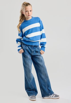 Suéter de rayas azul y blanco con cuello redondo, combinado con jeans de pierna ancha. Las zapatillas cuentan con acentos en gris y blanco. Textura suave.