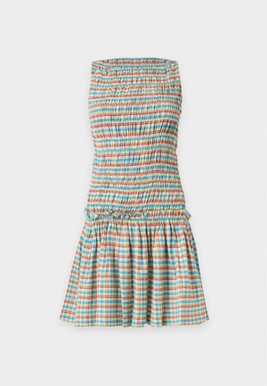 Kvinde iført en ærmeløs, flerfarvet ternet kjole, der holder en lille vævet taske og bærer beige slip-in sandaler mod en ensfarvet baggrund.