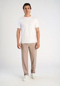 Mężczyzna stoi przodem, ubrany w białą koszulkę z krótkim rękawem, beżowe spodnie i białe sneakersy na jasnym, jednolitym tle.