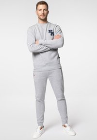 Szary sweatshirt i dopasowane dresy z miękkiego materiału, z małym granatowym logo na klatce piersiowej i zamkami błyskawicznymi na kostkach. Białe sneakersy dopełniają całość.