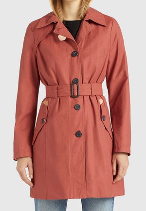 Woman wearing a rust-colored belted trench coat with black buttons over a black top and light blue jeans, shown from neck to mid-thigh.