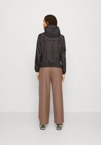 Zwarte lichte jas met een capuchon, elastische manchetten en taille; gecombineerd met een paar lange, bruine broek met wijde pijpen en sneakers. Achteraanzicht getoond.