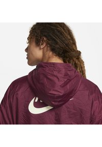 Bordo vetrovka s kapuco, ki ima na hrbtni strani velik bel Nike logo. Material izgleda gubano in lahkotno.