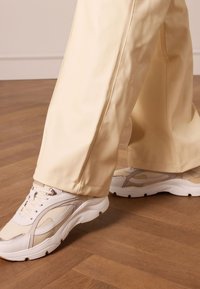 Lichtbeige sneakers met mesh- en lederen texturen; dikke witte zolen. Gecombineerd met crèmekleurige wijde broek, tentoongesteld op houten vloeren.