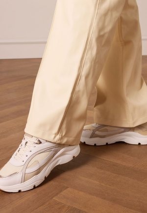 Light beige sneakers with mesh and leather textures; thick white soles. Paired with cream-colored wide-leg pants, displayed on wood flooring.