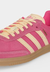 Ροζ αθλητικό παπούτσι Adidas Samba με κορδόνια και ρίγες σε εκρού, λαστιχένια σόλα και χρυσό λογότυπο "SAMBA" στο πλάι.