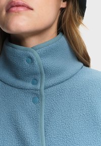 Gros plan sur une personne portant une veste polaire bleu clair à col montant avec trois boutons-pression fermés.