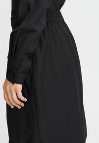 Schwarzes Kleid mit elastischem Taillenschneider, langen Ärmeln und Knopfmanschetten. Glatter Stoff, taillierte Passform, die zum Saum hin schmaler wird.