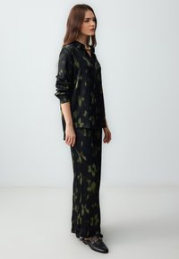 Zwarte en groene gemusterde, lichtgewicht stoffen broek en bijpassend shirt met een kraag, lange mouwen en een ronde onderrand. Platte zwarte schoenen.