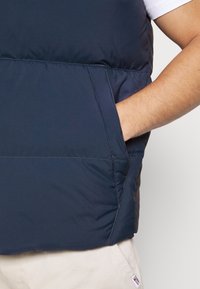 Mörkblå vadderad väst med slät textur och sidoficka, med kontrasterande sömmar. Bärs över en vit t-shirt och beige byxor.